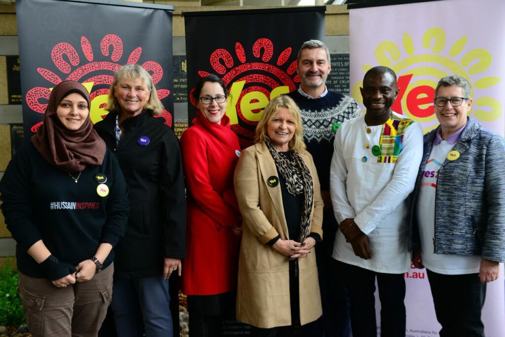 Photo of 7 people standing in front of 3 vertical banners with the branding of the 'YES' campaign.
